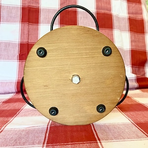 K&ouml;nnte beinhalten: Ein runder Servierteller aus Holz mit einem schwarzen Metallgriff. Das Tablett hat eine hellbraune Oberfl&auml;che und ist mit vier schwarzen Schrauben befestigt. In der Mitte befindet sich eine silberne Schraube. Der Hintergrund ist eine rot-wei&szlig; karierte Tischdecke.