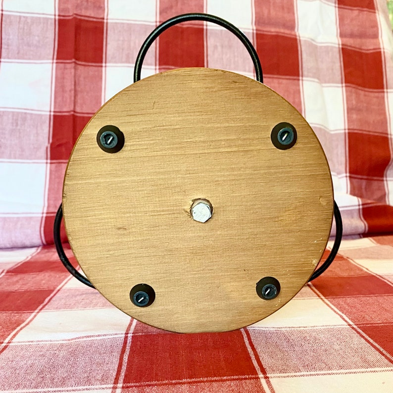K&ouml;nnte beinhalten: Ein runder Servierteller aus Holz mit einem schwarzen Metallgriff. Das Tablett hat eine hellbraune Oberfl&auml;che und ist mit vier schwarzen Schrauben befestigt. In der Mitte befindet sich eine silberne Schraube. Der Hintergrund ist eine rot-wei&szlig; karierte Tischdecke.