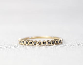 May include: A thin gold band ring featuring a series of small, round, black stones set within individual geometric, diamond-shaped metal frames that span the entire circumference of the band, creating a continuous, textured, and repeating pattern.