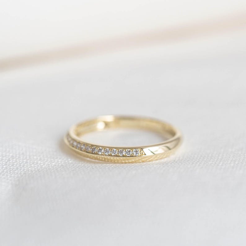 May include: A gold ring with a band of small, clear gemstones set along the top edge. The ring has a polished finish and sits on a white surface. The ring is a piece of jewelry.