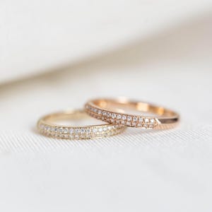Puede incluir: Dos anillos de oro, uno amarillo y otro oro rosa, cada uno adornado con una banda de peque&ntilde;os diamantes brillantes. Los anillos est&aacute;n sobre una superficie blanca texturizada. Probablemente son alianzas o anillos de compromiso.