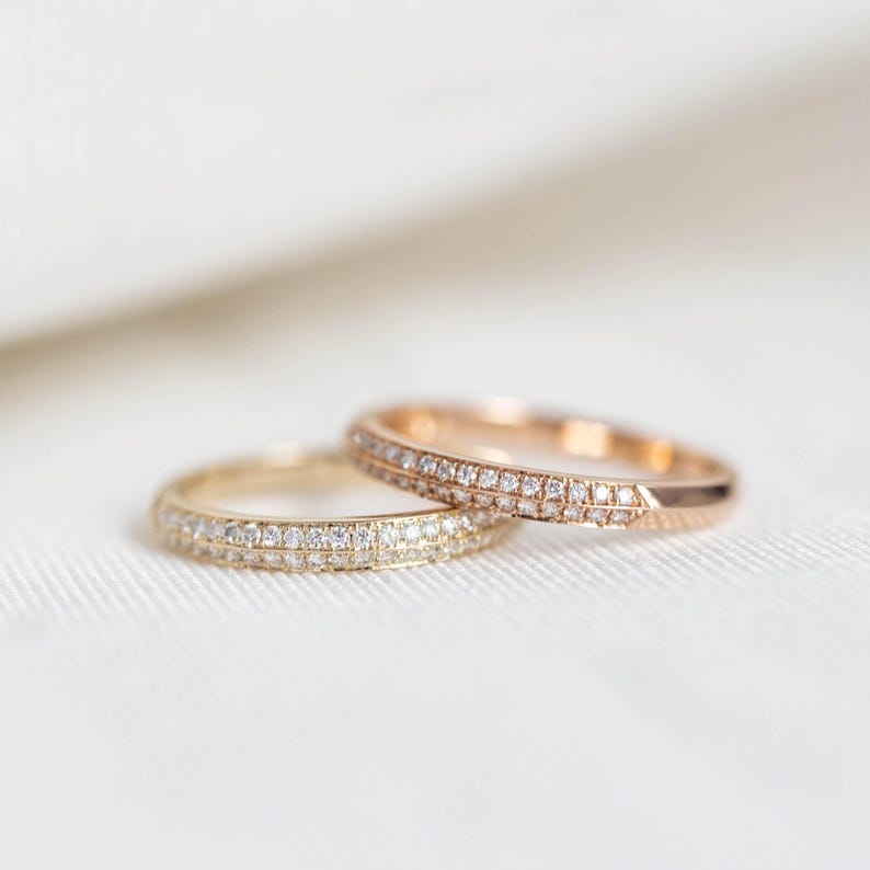 Puede incluir: Dos anillos de oro, uno amarillo y otro oro rosa, cada uno adornado con una banda de peque&ntilde;os diamantes brillantes. Los anillos est&aacute;n sobre una superficie blanca texturizada. Probablemente son alianzas o anillos de compromiso.