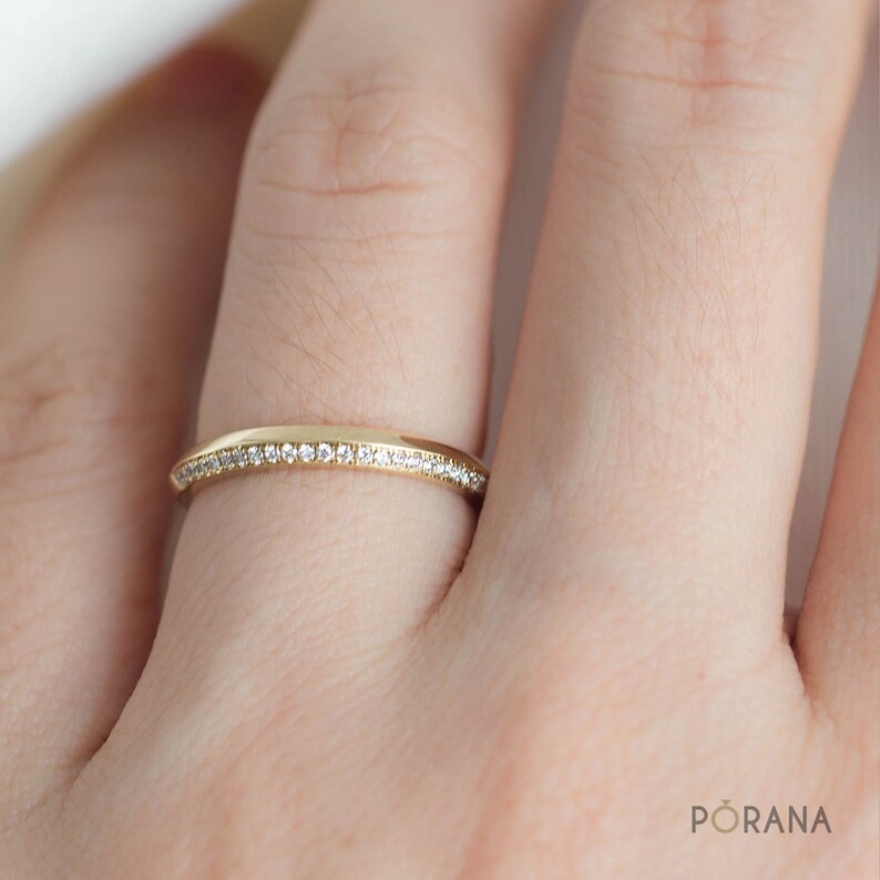 May include: A gold band ring with small, clear gemstones set along the top. The ring is on a finger, and the band has a smooth, polished finish. The ring is a delicate piece of jewelry.