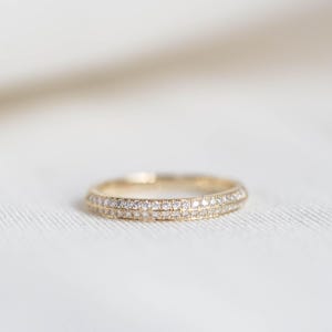 Puede incluir: Un anillo de banda de oro con dos filas de peque&ntilde;as piedras preciosas transparentes. El anillo se encuentra sobre un fondo blanco suave, resaltando su delicado dise&ntilde;o y brillo. Es una pieza de joyer&iacute;a.