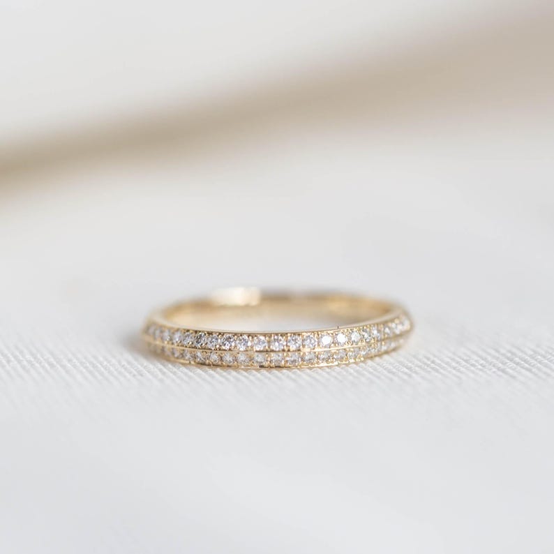 Puede incluir: Un anillo de banda de oro con dos filas de peque&ntilde;as piedras preciosas transparentes. El anillo se encuentra sobre un fondo blanco suave, resaltando su delicado dise&ntilde;o y brillo. Es una pieza de joyer&iacute;a.