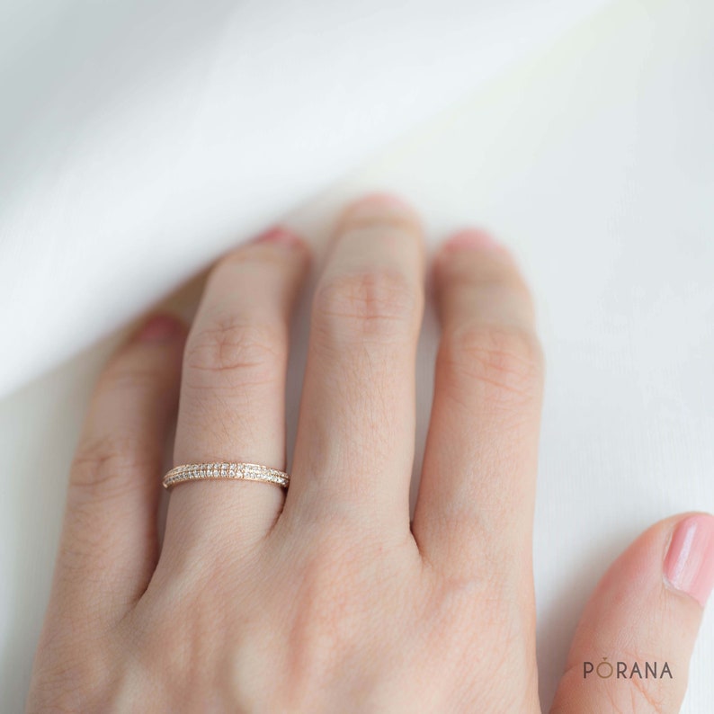 Puede incluir: Un anillo dorado con dos filas de peque&ntilde;as piedras transparentes. El anillo est&aacute; en el dedo de una persona, sobre un fondo blanco y suave. Es una joya delicada.