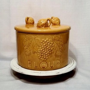 May include: A yellow ceramic flour canister with a grapevine design and the word "FLOUR" on the front. The canister has three decorative grapevine handles on the lid and sits on a white pedestal.