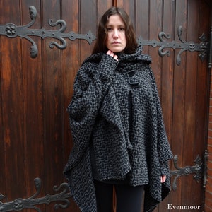 Könnte beinhalten: Ein schwarzer und grauer Kapuzenumhang mit geometrischem Muster. Der Umhang hat eine große Kapuze und ein langes, fließendes Design. Der Umhang wird von einer Person getragen, die vor einer Holztür steht.