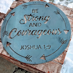May include: A round metal sign with a blue-grey finish. The sign has a cut-out design of arrows pointing in a circle around the words "Be Strong and Courageous" and the text "Joshua 1:9".