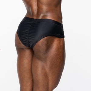 May include: Solid black underwear featuring a smooth, form-fitting fabric construction with a gathered center seam detail along the back. The garment has a low-rise waistband and a brief-style cut that provides full coverage across the rear.