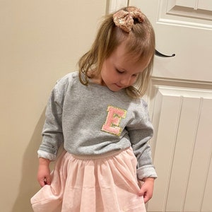 May include: A young child wearing a gray sweatshirt with a pink and gold letter "E" patch, a light pink tulle skirt, and black strappy sandals. A sparkly bow is in the child's hair.