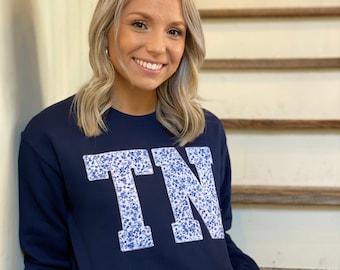 Könnte beinhalten: Ein marineblauer Sweatshirt mit einem weißen und blauen floralen "TN"-Motiv auf der Vorderseite.