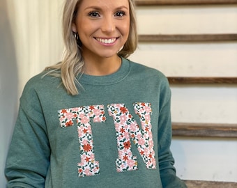 Könnte beinhalten: Ein grünes Sweatshirt mit einem floralen "TN"-Design. Das Sweatshirt hat eine lockere Passform und lange Ärmel.