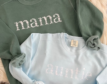 Op de afbeelding: Twee sweatshirts, een groene en een lichtblauwe, met de woorden "mama" en "auntie" geborduurd in wit op de voorkant. De borduurwerk is gemaakt van kleine witte bloemen.
