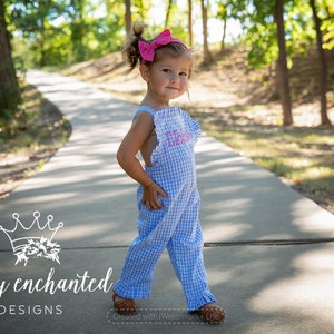 Puede incluir: Una niña pequeña con un mono de cuadros vichy azul y blanco con ribetes de volantes rosas y un lazo rosa en el pelo. El mono tiene el nombre "Ella Rose" bordado en rosa en la parte delantera.