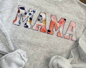 Op de afbeelding: Een grijs sweatshirt met het woord "MAMA" in grote, kleurrijke letters. De letters zijn omrand met zwart en gevuld met een bloemenpatroon.