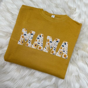Pu&ograve; includere: Una felpa gialla senape con la parola "MAMA" scritta in grandi lettere. Le lettere sono fatte di un tessuto bianco con un motivo floreale.