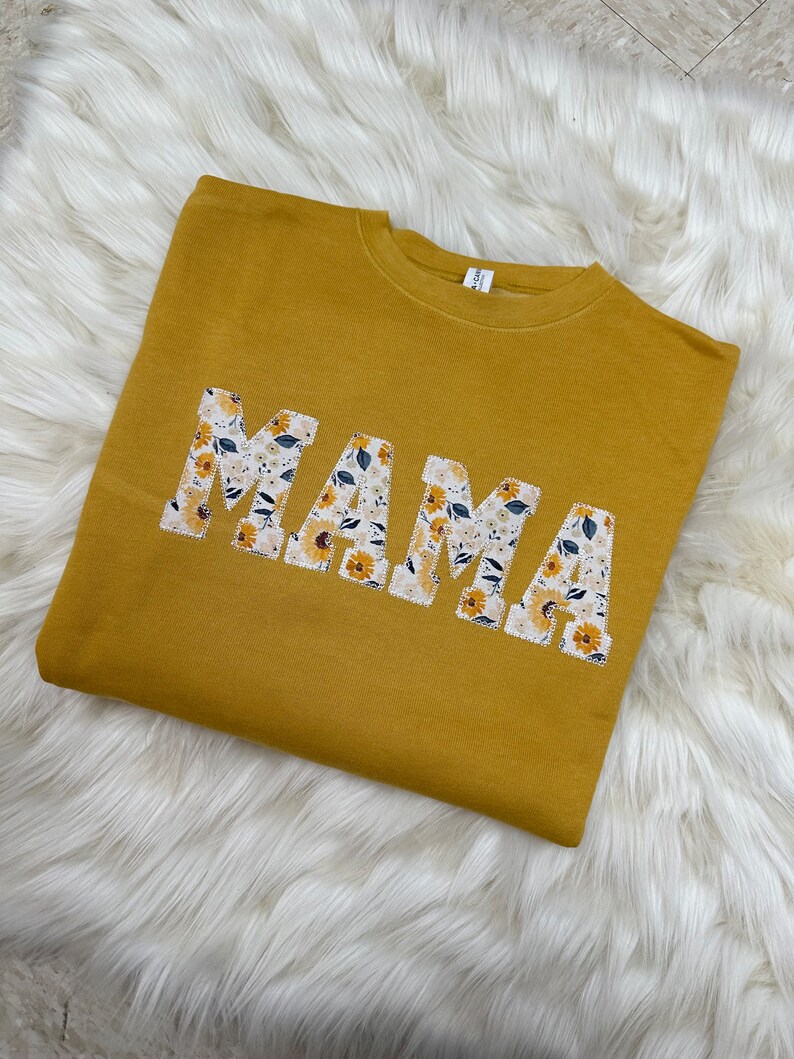 Pu&ograve; includere: Una felpa gialla senape con la parola "MAMA" scritta in grandi lettere. Le lettere sono fatte di un tessuto bianco con un motivo floreale.
