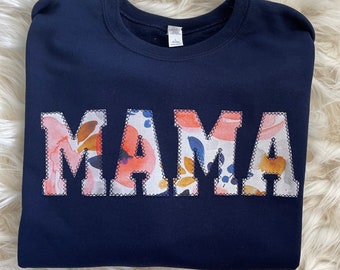Op de afbeelding: Marineblauw sweatshirt met het woord "MAMA" in letters met bloemenprint. De letters zijn omrand met een decoratieve steek. Het sweatshirt ligt op een witte, donzige ondergrond.