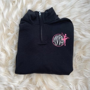 Puede incluir: Sudadera negra con cremallera de un cuarto con una silueta de bailarina rosa y iniciales monogramadas en un círculo.