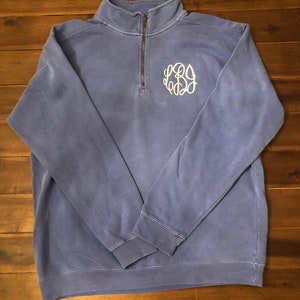 May include: A blue pullover sweatshirt with a quarter-zip neckline and a white monogram on the chest. The sweatshirt has long sleeves and a ribbed hem and cuffs. The fabric has a slightly faded appearance.