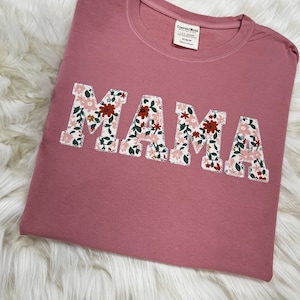 Könnte beinhalten: Ein pinkfarbenes T-Shirt mit dem Wort "MAMA" in großen weißen Buchstaben. Jeder Buchstabe ist mit einem Blumenmuster verziert.