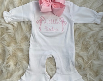 May include: White baby onesie with a pink bow and the text "Little Sister" embroidered on the front. The onesie has a ruffled bottom.