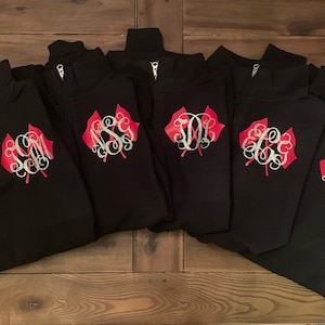 May include: Five black quarter-zip sweatshirts with red and silver embroidered flags and monograms. The monograms are "SM", "JM", "KG", "EC", and "LL".