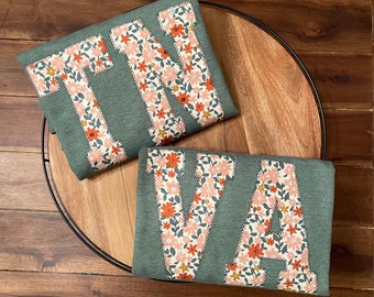 Könnte beinhalten: Zwei grüne Stoffartikel, möglicherweise Kissen oder Aufnäher, jeweils mit einem großen Buchstaben mit Blumenmuster. Einer zeigt "TN" und der andere "VA". Die Buchstaben sind im Patchwork-Stil mit floralem Design.