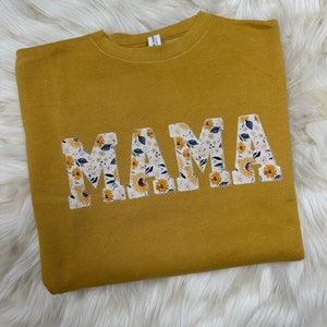 Pu&ograve; includere: Una felpa gialla senape con la parola "MAMA" in grandi lettere bianche. Le lettere sono decorate con un motivo floreale.
