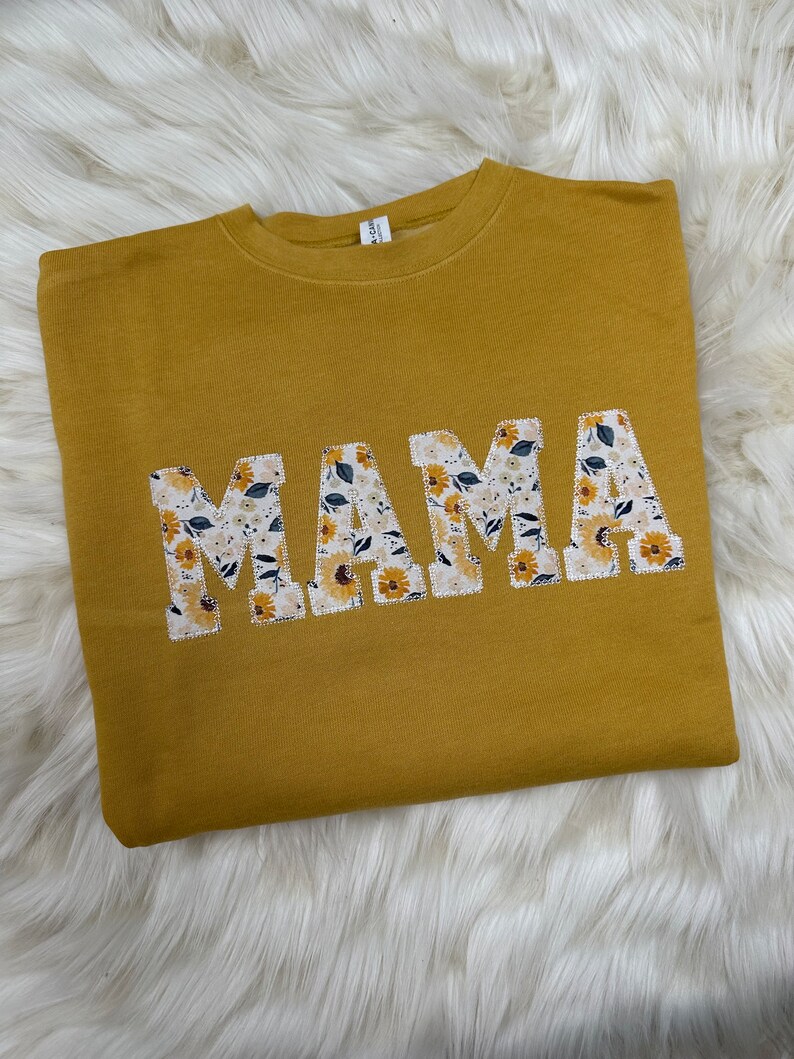 Pu&ograve; includere: Una felpa gialla senape con la parola "MAMA" in grandi lettere bianche. Le lettere sono decorate con un motivo floreale.