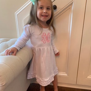 Peut inclure: Une jeune fille portant une robe blanche à manches longues avec un monogramme rose sur le devant. La robe a un ourlet à volants et la fille porte un nœud vert clair dans les cheveux.