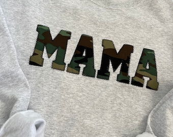May include: Gray sweatshirt with the word "MAMA" in large, black letters with a camouflage pattern.