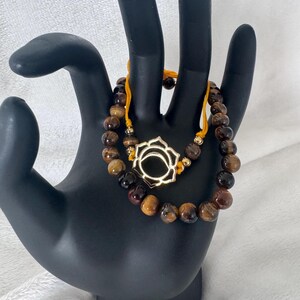 May include: Two beaded bracelets featuring tiger eye stones and a gold-coloured lotus flower charm. The bracelets are strung on a yellow cord and are displayed on a black hand.