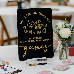 May include: A black sign with gold text that reads "Don't feel like dancing? No worries! Grab a friend and play some games." The sign is on a white table with board games, dice, and playing cards. A small vase of white roses is also on the table.