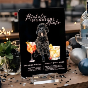 May include: A black sign with the text "Mabel's Signature Drinks" and illustrations of two cocktails and a black dog. The sign lists the ingredients for each drink: Devotion Potion and Apple-Y Ever After. The sign is on a wooden table.