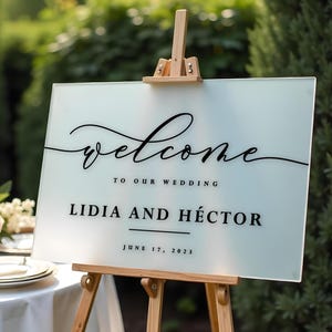 May include: White frosted welcome sign with black calligraphy lettering on an easel. The sign reads "Welcome to our wedding Lidia and Héctor June 17, 2023".