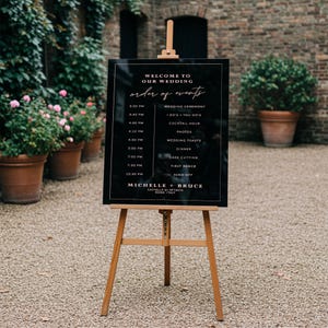 May include: Black wedding sign with a white frame and gold lettering. The sign lists the order of events for a wedding, including the ceremony, cocktail hour, dinner, and first dance. The sign also includes the names Michelle and Bruce, the location Castello di Petrata, and the city Rome, Italy.