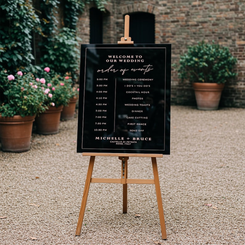 May include: Black wedding sign with a white frame and gold lettering. The sign lists the order of events for a wedding, including the ceremony, cocktail hour, dinner, and first dance. The sign also includes the names Michelle and Bruce, the location Castello di Petrata, and the city Rome, Italy.