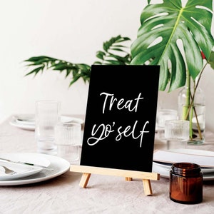 K&ouml;nnte beinhalten: Ein schwarzes Schiefertafel-Staffeleischild mit wei&szlig;em Text, der "Treat yo'self" lautet. Das Schild steht auf einem Tisch mit einer braunen Tischdecke und einem Glasgef&auml;&szlig; mit braunem Deckel.