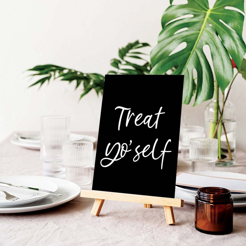 K&ouml;nnte beinhalten: Ein schwarzes Schiefertafel-Staffeleischild mit wei&szlig;em Text, der "Treat yo'self" lautet. Das Schild steht auf einem Tisch mit einer braunen Tischdecke und einem Glasgef&auml;&szlig; mit braunem Deckel.