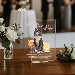 May include: Clear acrylic sign featuring a gray and white cat illustration. The sign reads "Natred's Signature Drinks" and lists two cocktail recipes: "Devotion Potion" and "Apple-Y Ever After". The sign is displayed on a wooden table with floral arrangements.