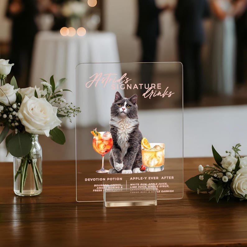 May include: Clear acrylic sign featuring a gray and white cat illustration. The sign reads "Natred's Signature Drinks" and lists two cocktail recipes: "Devotion Potion" and "Apple-Y Ever After". The sign is displayed on a wooden table with floral arrangements.