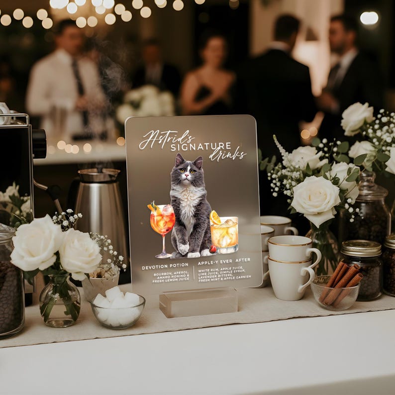 May include: A sign for "Astrid's Signature Drinks" featuring a gray cat illustration with two cocktail glasses. The sign is on a table with white roses, coffee beans, sugar cubes, and other bar accessories.