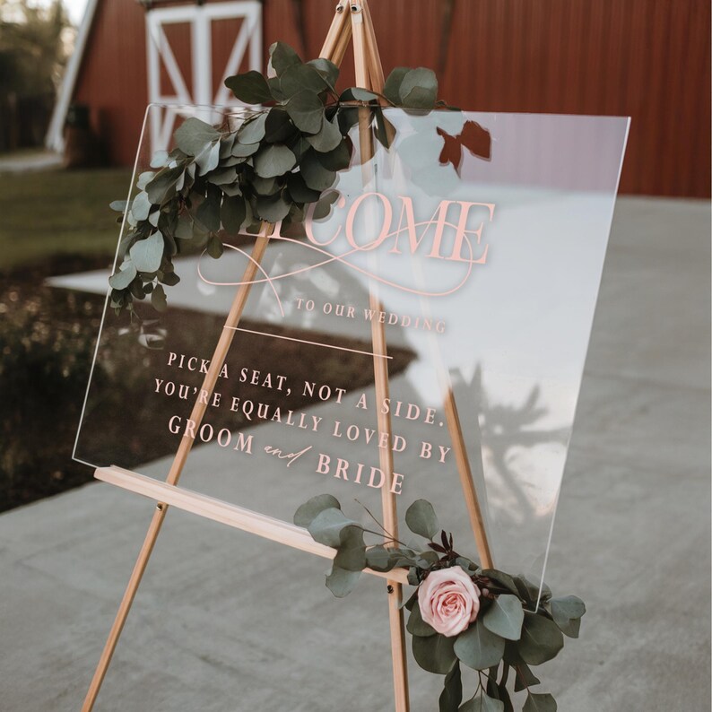 Pu&ograve; includere: Cartello di benvenuto per matrimoni in acrilico trasparente con scritta color oro rosa e decorazioni floreali. Il cartello &egrave; esposto su un cavalletto di legno e presenta il testo "WELCOME TO OUR WEDDING" e un messaggio sui posti a sedere.