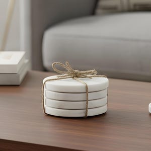May include: A stack of four white marble coasters tied with natural twine. The coasters are round and have a smooth surface. The twine is tied in a bow at the top. The coasters are on a brown wooden table.
