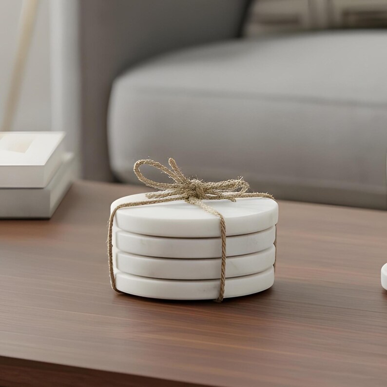 May include: A stack of four white marble coasters tied with natural twine. The coasters are round and have a smooth surface. The twine is tied in a bow at the top. The coasters are on a brown wooden table.