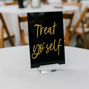 K&ouml;nnte beinhalten: Schwarzes Acrylschild mit goldener Aufschrift "Treat yo' self" auf einem klaren Acrylst&auml;nder.