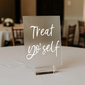 K&ouml;nnte beinhalten: Ein klares Acrylschild mit einer wei&szlig;en Aufschrift, die "Treat yo' self" lautet, auf einem klaren Acrylst&auml;nder.