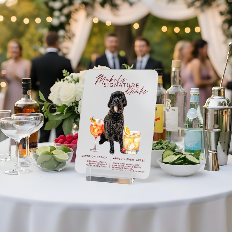 May include: A white sign with the text "Mabel's Signature Drinks" and a watercolor illustration of a black dog. The sign lists two cocktail recipes: "Devotion Potion" and "Apple-Y Ever After". The sign is displayed on a white tablecloth with cocktail glasses and bottles.